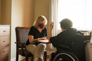 Healthcare worker wearing scrubs, gloves and a red fabric face mask sits across from a patient on oxygen sitting in a wheelchair during a speech therapy session in the COVID-19 pandemic.