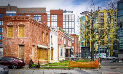 Row of stone buildings with boarded up windows, with new tall buildings all around.