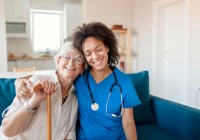 A nurse in blue scrubs has her right arm around the shoulders of an older woman holding a wooden cane. Both are seated on a blue couch.