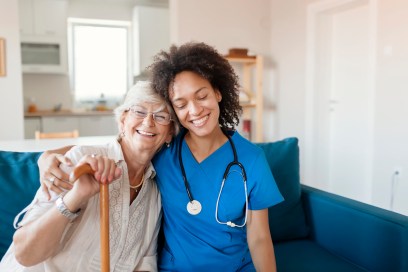 A nurse in blue scrubs has her right arm around the shoulders of an older woman holding a wooden cane. Both are seated on a blue couch.