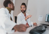 Dark skinned people wearing white coats talking in front of a computer