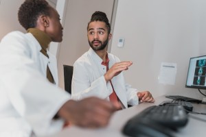 Dark skinned woman and dark skinned man wearing white coats talking in front of a computer