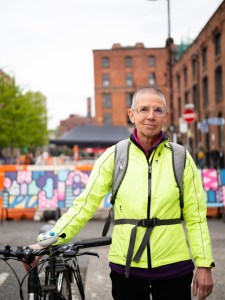 Older person with short grey hair wearing a bright yellow jacket and standing with their hand on a bicycle.