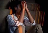 Person with dark skin sitting on the ground with their elbows on knees and hands on their face.