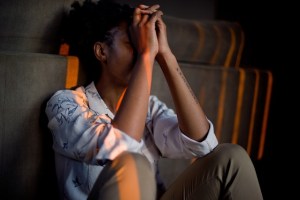 Person with dark skin sitting on the ground with their elbows on knees and hands on their face. 