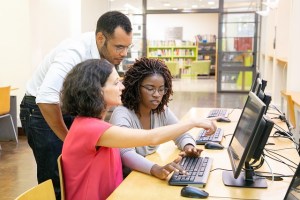 Three university students, one dark-skinned woman, one light-skinned woman, and one dark-skinned man, talking and working at a computer in a library